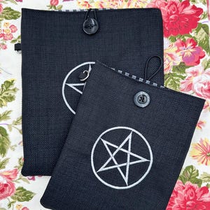 May include: Two black pouches with white embroidered symbols. One pouch has a circle with a crescent moon and a V shape inside. The other pouch has a white pentagram symbol. Both pouches have a button closure and a loop.