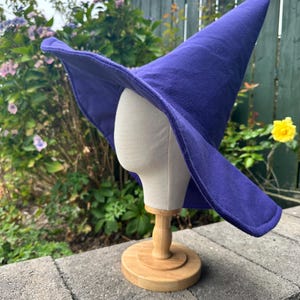 May include: A vibrant purple witch hat with a wide, flexible brim and a tall, pointed crown. The hat is constructed from a solid-colored fabric and is displayed on a neutral-toned mannequin head stand with a circular wooden base.