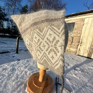 Può includere: Un cappello di lana bianco e grigio con un motivo geometrico. Il cappello ha un lungo fiocco grigio ed è esposto su un supporto in legno su uno sfondo innevato.