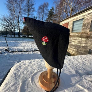 Peut inclure: Un chapeau noir orné d'une broderie de champignon rouge et blanc. Le chapeau est présenté sur un support en bois, avec un cordon noir. L'arrière-plan montre un paysage enneigé avec des arbres et une structure en bois.