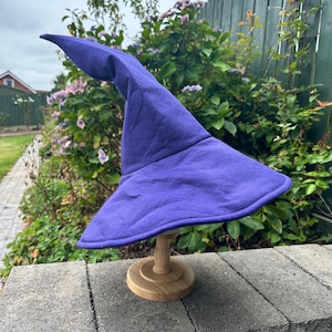 May include: A purple pointed hat with a wide, flexible brim. The hat features a tall, slightly curved cone shape and is constructed from a solid-colored fabric. It is displayed on a small, light-colored wooden stand with a circular base.