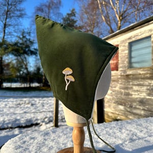 May include: A dark green bonnet with a white lining and an embroidered mushroom motif. The bonnet is displayed on a wooden stand. The bonnet has drawstrings. The background is snowy.