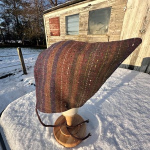 May include: A striped knit hat in shades of brown, purple, and green, displayed on a wooden stand. The hat has a pointed end and vertical stripes. The hat is displayed on a wooden stand with a round base and a white head form.