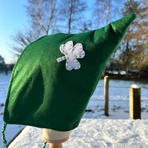 Peut inclure: Un chapeau de lutin en feutre vert avec un trèfle blanc cousu. Le chapeau a une pointe et un cordon tressé vert et blanc. Le chapeau est présenté sur un support en bois dans un cadre extérieur enneigé.