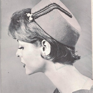 May include: A vintage-style hat with a square crown and a decorative beaded trim. The hat is a light gray color and features a small cluster of white beads on the side. The image is a black and white photograph.