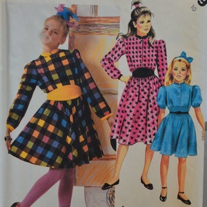 Puede incluir: Tres chicas vistiendo vestidos. La chica de la izquierda lleva un vestido de cuadros con un cinturón amarillo. La chica del medio lleva un vestido rosa con un cinturón negro. La chica de la derecha lleva un vestido azul con un cuello blanco.