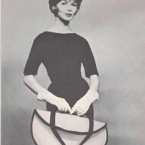 May include: A black dress with short sleeves and a boat neckline. The model is wearing white gloves and holding a large, semi-circular handbag with a cream-colored body and black straps. The image is in black and white.