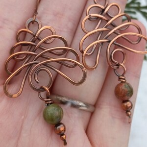 May include: A pair of copper wire earrings with a delicate, intricate design. Each earring features a dangling green and brown stone.