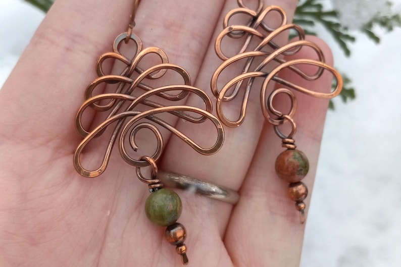 May include: A pair of copper wire earrings with a delicate, intricate design. Each earring features a dangling green and brown stone.