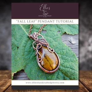 Puede incluir: Un colgante de cobre hecho a mano con una piedra de ojo de tigre en forma de lágrima, exhibido sobre una hoja verde. El colgante forma parte de un tutorial titulado "Fall Leaf" y se muestra sobre una superficie de madera.