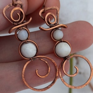 May include: A pair of copper wire earrings with white beads, shaped like snowmen. The earrings have a spiral design at the bottom.