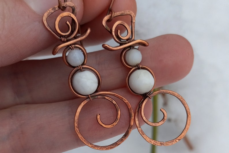 May include: A pair of copper wire earrings with white beads, shaped like snowmen. The earrings have a spiral design at the bottom.