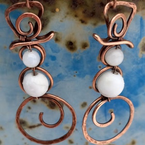 May include: A pair of copper wire snowman earrings with white beads. The earrings are handmade and feature a spiral design.