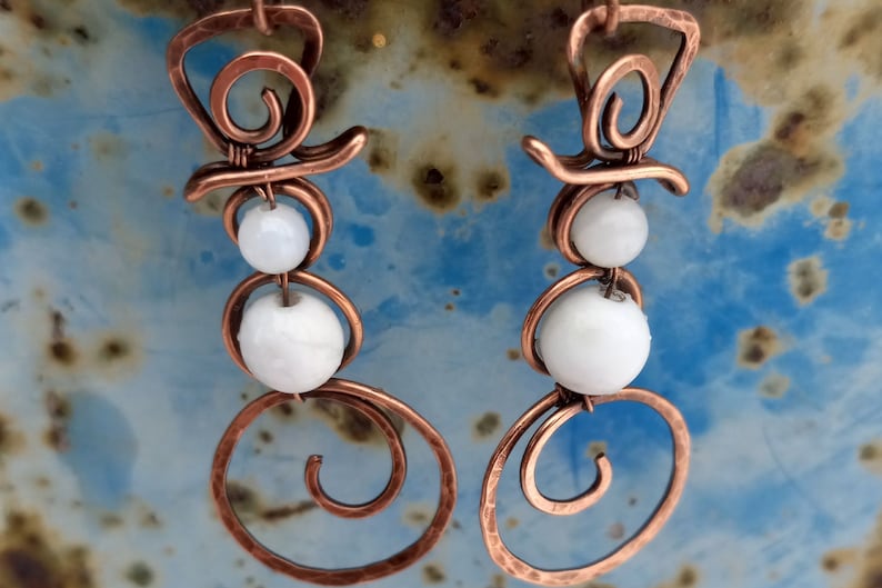 May include: A pair of copper wire snowman earrings with white beads. The earrings are handmade and feature a spiral design.