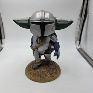 May include: A silver and blue figurine of a character from the Star Wars franchise, standing on a brown, circular base. The figure is wearing a helmet with large, green-tipped ears, armor, and a utility belt. The figure is holding a small weapon.