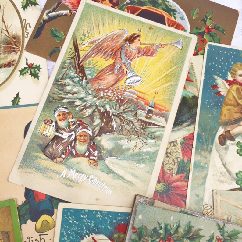 May include: A collection of vintage Christmas postcards featuring a variety of festive scenes. One postcard depicts an angel with wings blowing a trumpet, another shows two figures with lanterns in a snowy forest, and a third features a child holding a Christmas tree. The postcards are printed on thick paper with colourful illustrations and gold accents.