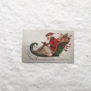 May include: Vintage Christmas postcard featuring Santa Claus in a green sleigh with a red coat and white beard, pulling a small child with a Christmas tree. The text "Merry Xmas" is written in cursive on the postcard.