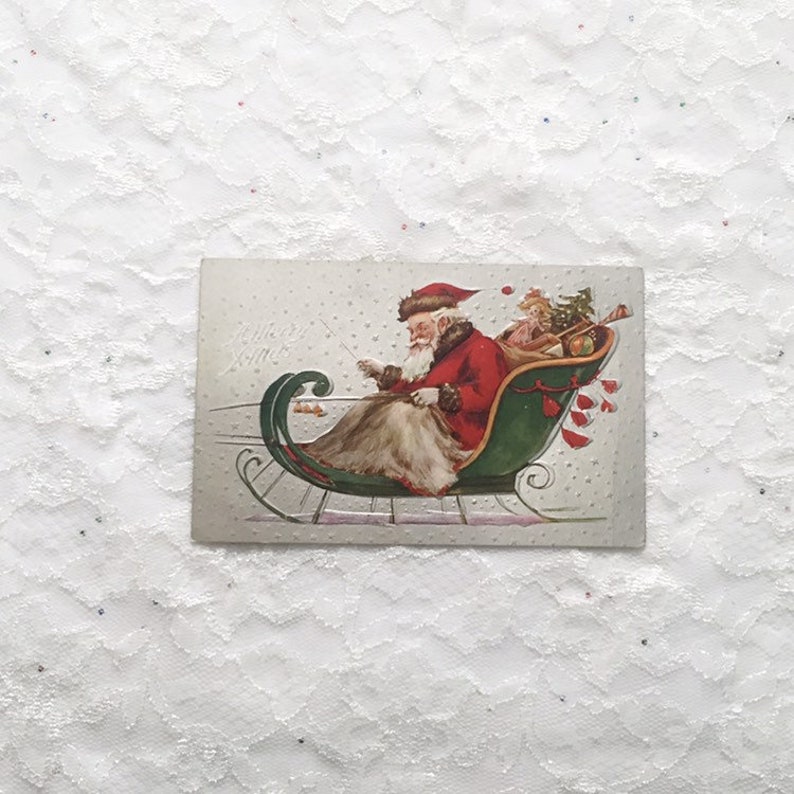 May include: Vintage Christmas postcard featuring Santa Claus in a green sleigh with a red coat and white beard, pulling a small child with a Christmas tree. The text "Merry Xmas" is written in cursive on the postcard.