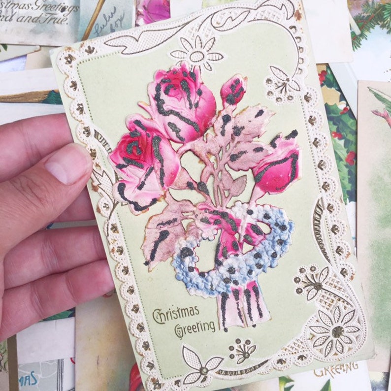 May include: Vintage Christmas greeting card featuring a bouquet of pink roses and a blue and white ribbon. The card has a lace border and the text "Christmas Greeting" is printed on the front.