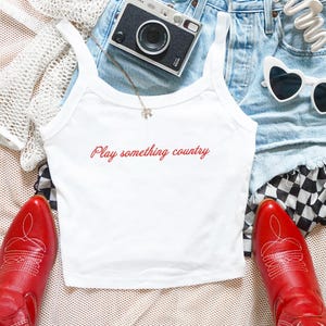 May include: A white tank top with the red text Play something country printed on the front, paired with light blue denim shorts, a black instant camera, white heart-shaped sunglasses, and bright red cowboy boots with white stitching details.