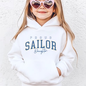 Könnte beinhalten: Weißer Kapuzenpullover mit dem Text "PROUD SAILOR Daughter" in blauer und gelber Schrift.