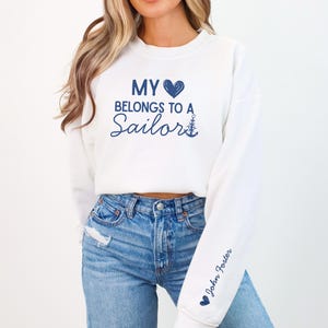 May include: White sweatshirt with the text "MY BELONGS TO A Sailor" in navy blue script. The sleeve has the name "John Foster" in navy blue script. The sweatshirt is paired with blue jeans.