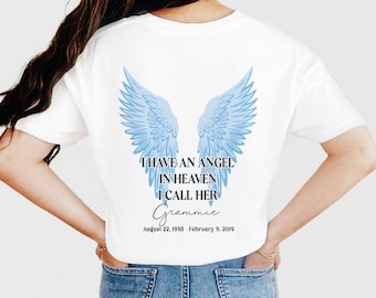 May include: White t-shirt with blue angel wings graphic and the text "I HAVE AN ANGEL IN HEAVEN I CALL HER Grammie August 22, 1938 - February 9, 2019". The shirt is a casual style.