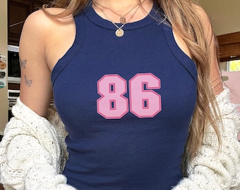 May include: A navy blue tank top with the number "86" in pink block letters. The top has a round neckline and a racerback style. The image shows a close-up of the top, highlighting the color and design.