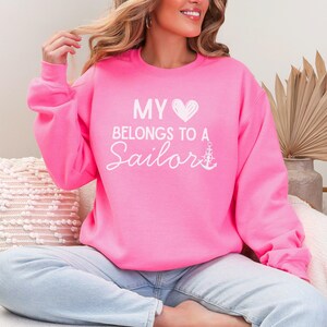 May include: Bright pink sweatshirt with the text "MY HEART BELONGS TO A Sailor" in white script. The design includes a white heart and anchor. The sweatshirt is a crew neck style.