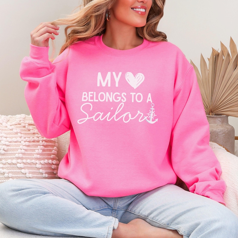 May include: Bright pink sweatshirt with the text "MY HEART BELONGS TO A Sailor" in white script. The design includes a white heart and anchor. The sweatshirt is a crew neck style.