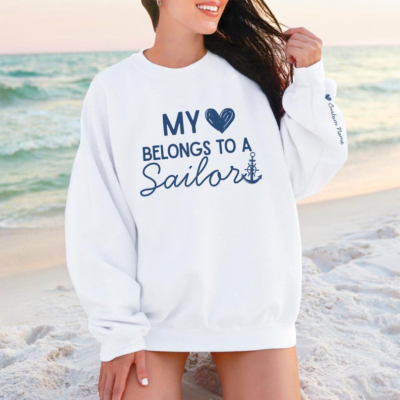 May include: White sweatshirt with the text "MY HEART BELONGS TO A Sailor" in navy blue. The design includes a navy blue heart and an anchor. The sleeve has the text "Custom Name" in navy blue.