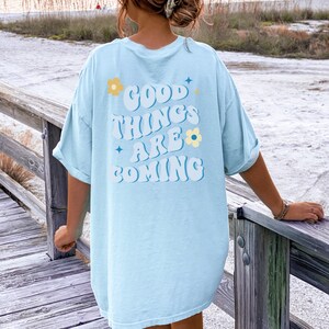 Puede incluir: Camiseta azul claro con el texto "GOOD THINGS ARE COMING" en blanco y azul, decorada con flores y estrellas. La camiseta es de corte holgado y tiene las mangas dobladas.