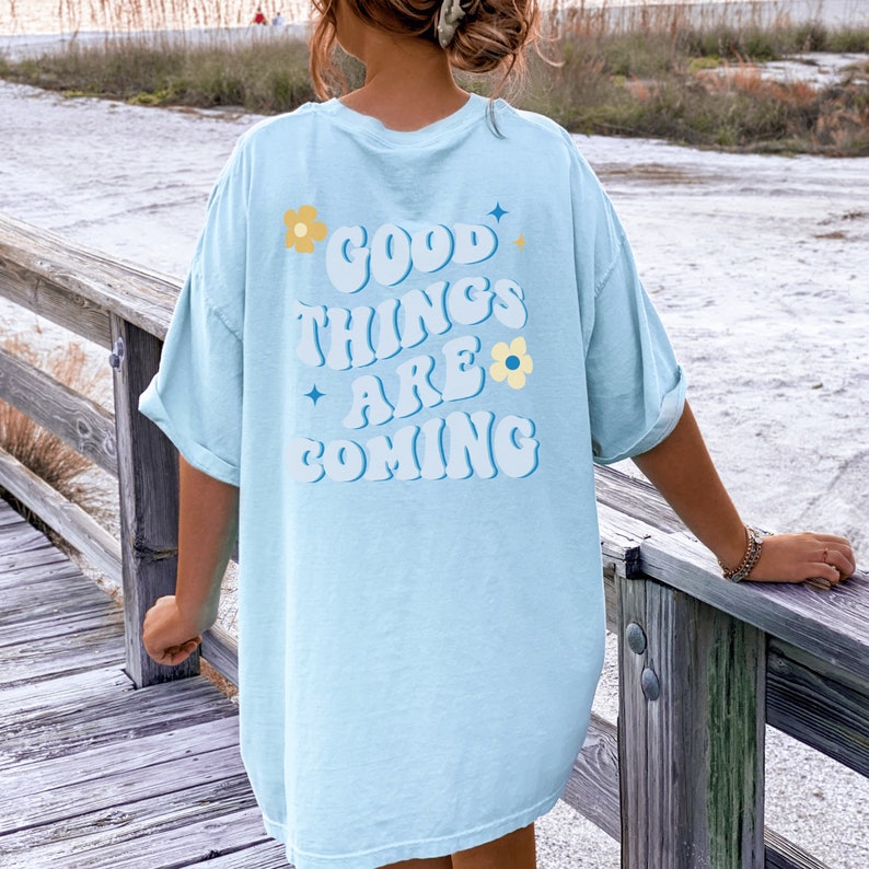 Puede incluir: Camiseta azul claro con el texto "GOOD THINGS ARE COMING" en blanco y azul, decorada con flores y estrellas. La camiseta es de corte holgado y tiene las mangas dobladas.
