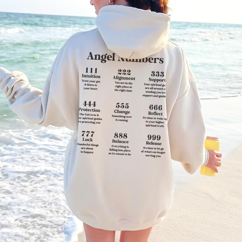 May include: Cream-colored hoodie with the words "Angel Numbers" printed on the back, along with various number sequences and their meanings. The hoodie is worn on a beach with the ocean in the background.