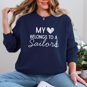 May include: Navy blue sweatshirt with the text "MY HEART BELONGS TO A Sailor" in white script. The design includes a white heart and an anchor. The sweatshirt is a crew neck style.