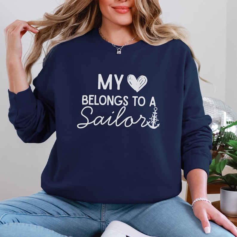May include: Navy blue sweatshirt with the text "MY HEART BELONGS TO A Sailor" in white script. The design includes a white heart and an anchor. The sweatshirt is a crew neck style.