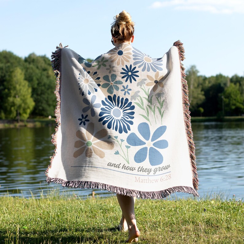May include: A beige throw blanket with a floral design in shades of blue and tan. The blanket has a fringed edge and the text "and how they grow Matthew 6:28". The blanket is being held up by a person.