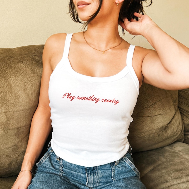 May include: A white ribbed tank top featuring the text Play something country in red cursive script across the chest. The garment has a scoop neckline and thick shoulder straps, paired with blue denim jeans.