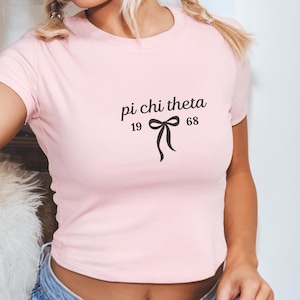 May include: A light pink crop top with the text "pi chi theta" above a bow graphic and the numbers "19" and "68". The shirt has short sleeves and a crew neck.