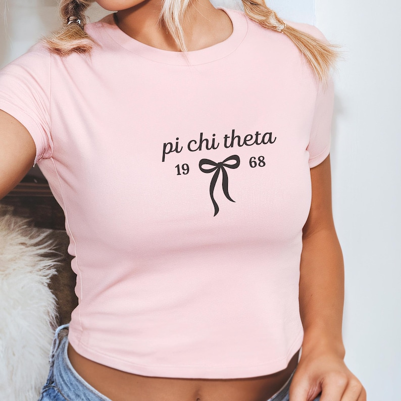 May include: A light pink crop top with the text "pi chi theta" above a bow graphic and the numbers "19" and "68". The shirt has short sleeves and a crew neck.