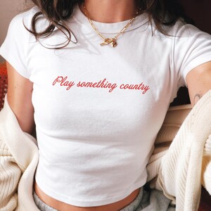 May include: A white short-sleeved t-shirt featuring the text Play something country printed in a red cursive font across the chest. The shirt has a crew neckline and is worn with a gold-toned chain necklace and a cream-colored cardigan.