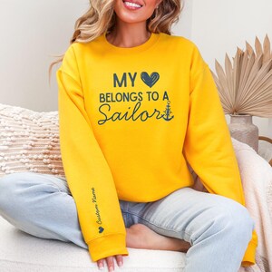May include: Mustard yellow sweatshirt with the text "MY 💙 BELONGS TO A Sailor" in navy blue. An anchor graphic is next to the word "Sailor". The left sleeve has the text "Custom Name" in navy blue with a small heart graphic.