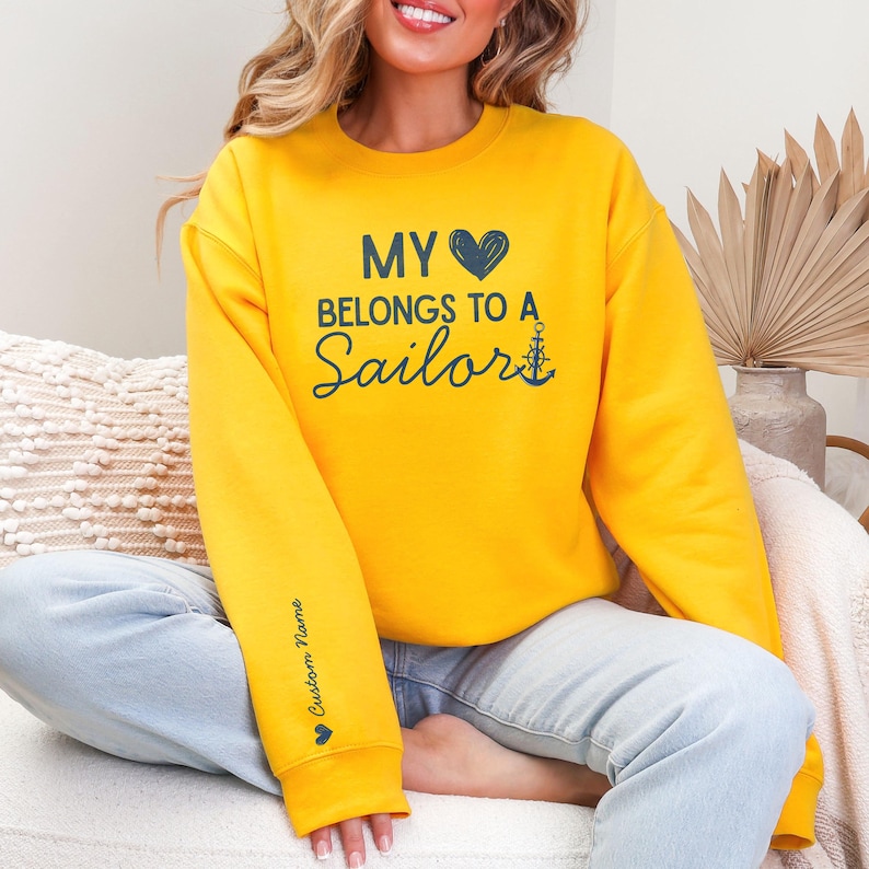 May include: Mustard yellow sweatshirt with the text "MY 💙 BELONGS TO A Sailor" in navy blue. An anchor graphic is next to the word "Sailor". The left sleeve has the text "Custom Name" in navy blue with a small heart graphic.