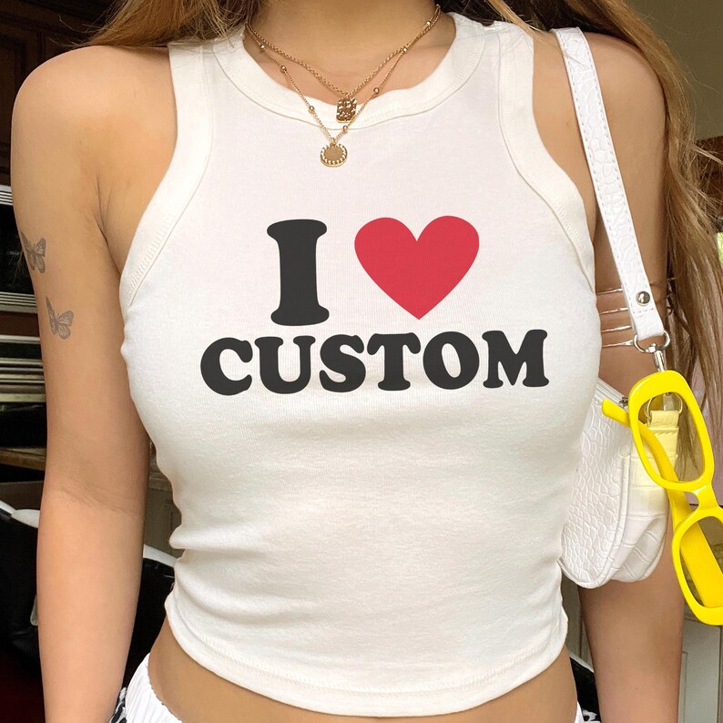 May include: White tank top with the text "I &hearts; CUSTOM" in black and red. The top is a sleeveless, ribbed style. A white purse and yellow sunglasses are visible.