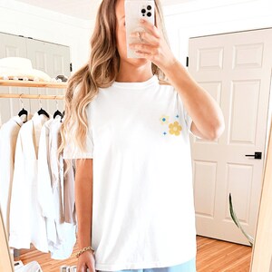 Puede incluir: Camiseta blanca con un peque&ntilde;o dise&ntilde;o floral en la parte superior izquierda del pecho. El dise&ntilde;o presenta una flor amarilla, una flor azul y dos formas azules en forma de estrella. La camiseta tiene mangas cortas y cuello redondo.