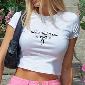 May include: White crop top with the text "delta alpha chi" in black script, along with the numbers "19" and "53" and a bow graphic. The top is short-sleeved and has a fitted silhouette.