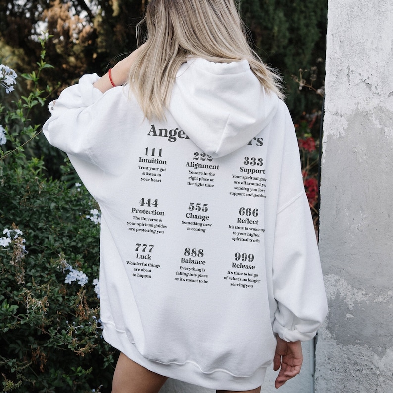 May include: White oversized hoodie with the text "Angel Numbers" and various number sequences with corresponding affirmations. The hoodie has a drawstring hood and long sleeves. The text is printed in black.