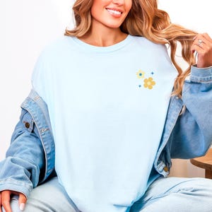 Puede incluir: Camiseta azul claro con un peque&ntilde;o dise&ntilde;o floral en amarillo y azul. La camiseta es de material suave y tiene un corte relajado. El dise&ntilde;o floral incluye dos flores y tres peque&ntilde;as explosiones estelares.