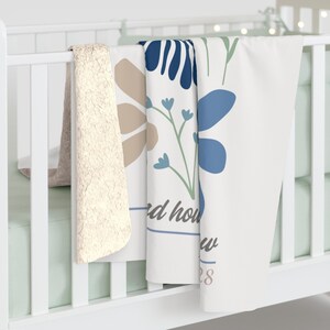 May include: A white baby blanket with a floral design in blue and tan, draped over a crib rail. The blanket features the text "d how w 28" in a script font. A cream-colored sherpa blanket is also visible.