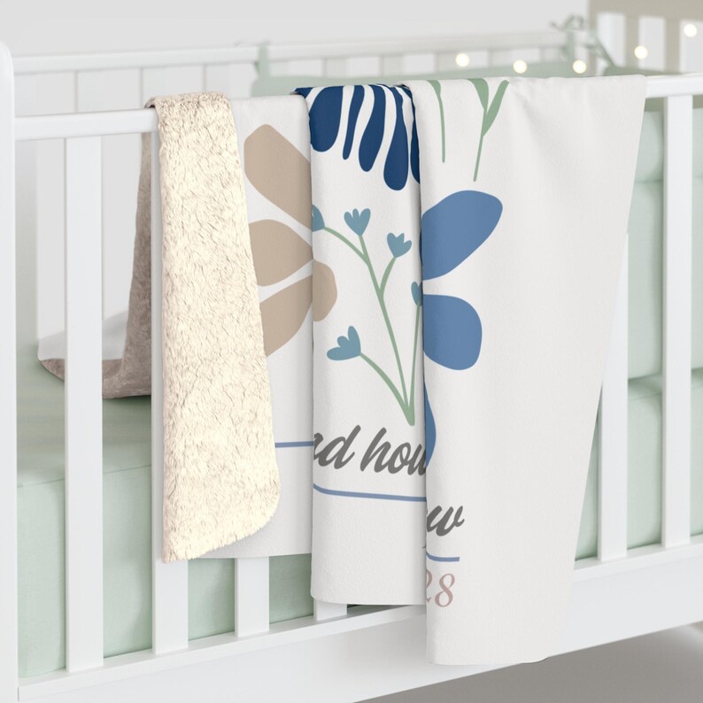 May include: A white baby blanket with a floral design in blue and tan, draped over a crib rail. The blanket features the text "d how w 28" in a script font. A cream-colored sherpa blanket is also visible.